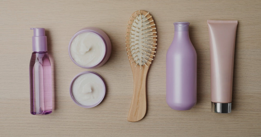 Flat lay of salon hair care products on a wooden surface: a hair oil, two jars of hair mask, a wooden paddle brush, a shampoo bottle and a conditioner tube.