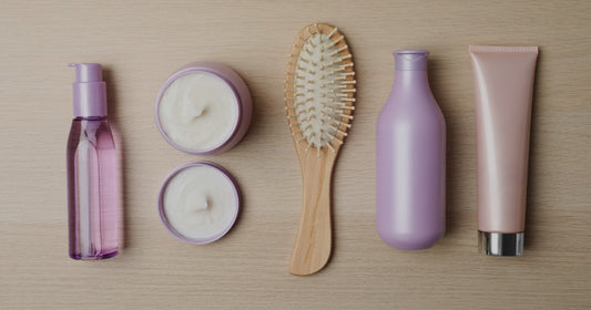Flat lay of salon hair care products on a wooden surface: a hair oil, two jars of hair mask, a wooden paddle brush, a shampoo bottle and a conditioner tube.